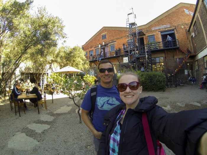 Selfie de Bruna e Diego na frente do Arts on Main, fábrica antiga de tijolos a vista onde hoje ocorre uma feira de comida e artesanato.
