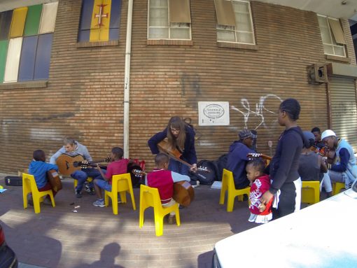 4 crianças tendo aulas de violão em uma calçada de Maboneng sentadas em cadeiras amarelas sob um fundo de tijolos aparentes.
